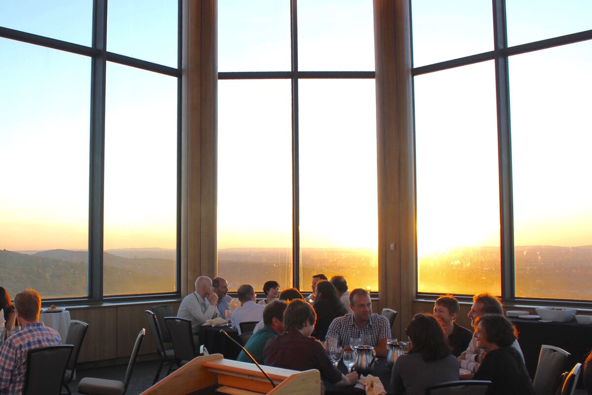 Attendees at dinner tables in front of tall windows at sunset